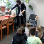 Na zdjęciu widać stojącą Panią Małgorzatę Swędrowską i siedzącą na dywanie grupę dzieci.